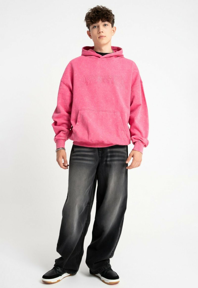 Różowa bluza z kapturem z kieszenią kangurką, z teksturowanym logo. Połączona z luźnymi czarnymi dżinsami i czarnymi trampkami z białymi podeszwami.