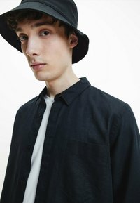 Camicia nera con bottoni, dotata di taschino sul petto, in tessuto leggero e con colletto, indossata sopra una t-shirt bianca, abbinata a un cappello a secchiello nero.