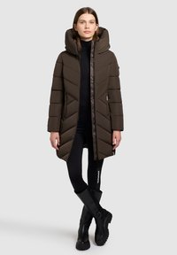 Cappotto imbottito marrone con cappuccio, caratterizzato da cuciture a chevron e chiusura zip sul davanti. La modella indossa leggings neri e stivali neri al ginocchio.