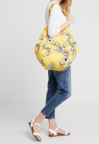 Borsa a tracolla floreale gialla con stampe nere di fiori, grande forma, tessuto resistente e lunghe tracolle per il trasporto a spalla, abbinata a jeans blu.