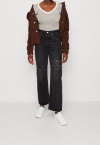 Schwarze hochgeschnittene Jeans mit angeschnittenen Details, kombiniert mit einem gestreiften Kurzarmoberteil und einer braunen Fleecejacke, abgerundet mit weißen Sneakers.