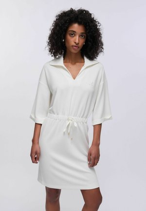 Ženska s kodrastimi lasmi, oblečena v belo obleko dolžine do kolen z zavezanim pasom in V-izrezom, stoji pred enobarvnim ozadjem.