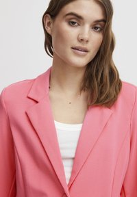 Rozā žakete ar iegrieztiem atlokiem un brīvu piegriezumu, izgatavota no viegla materiāla. M�ēģināta virs baltas topiņa, kas raksturojas ar gludu tekstūru.