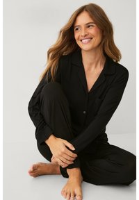 Ensemble de pyjama noir en tissu doux, comprenant une chemise à boutons avec une poche et un pantalon à coupe décontractée. Modèle assis pieds nus sur une surface grise.