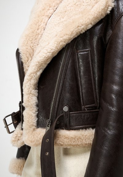 Jachetă din piele maro cu guler din blană bej, având un fermoar în față, detalii cu cataramă pe curea și cusături orizontale pe buzunare.