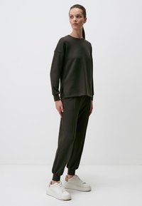 Dunkelgrüner, leichter Sweatshirt mit langen Ärmeln und seitlichen Schlitzen, kombiniert mit passenden Jogginghosen und weißen Turnschuhen. Einfaches Design, minimalistischer Stil.