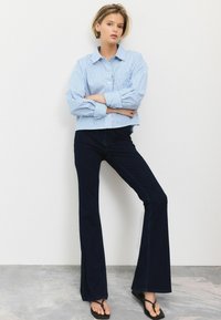 Gestreepte blauwe en witte cropped button-up shirt gecombineerd met donkere flared jeans en zwarte teenslippers, op een effen achtergrond.