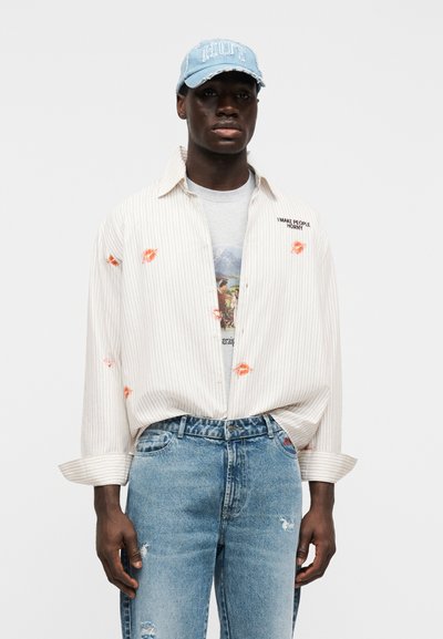 Ριγέ πουκάμισο σε μπεζ απόχρωση με πορτοκαλί αποτυπώματα φιλιών, φορεμένο πάνω από ένα γκρι t-shirt με γραφικά. Ανοιχτό μπλε σκισμένο καπέλο και ξεθωριασμένο μπλε τζιν.