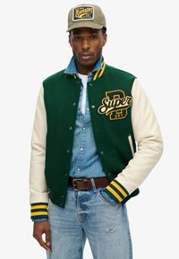 Chaqueta varsity verde y crema con puños a rayas amarillas, que presenta un gran logo de "Super". Lleva puesta sobre una camisa de denim y jeans azules.