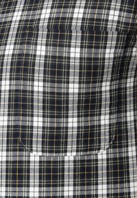 Kortärmad skjorta i ett rutmönster med svart, vitt och guld. Har en ficka och ett slätt, texturerat tyg.