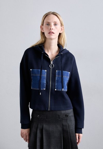 Sudadera corta de color navy con cremallera frontal, que cuenta con dos bolsillos de parche de mezclilla y cordones. Se lleva con una falda de cuadros con pliegues.