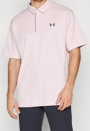 Mann trägt hellrosa Kurzarm-Poloshirt mit schwarzem Under Armour-Logo und dunkler Hose, steht vor einfarbigem Hintergrund.