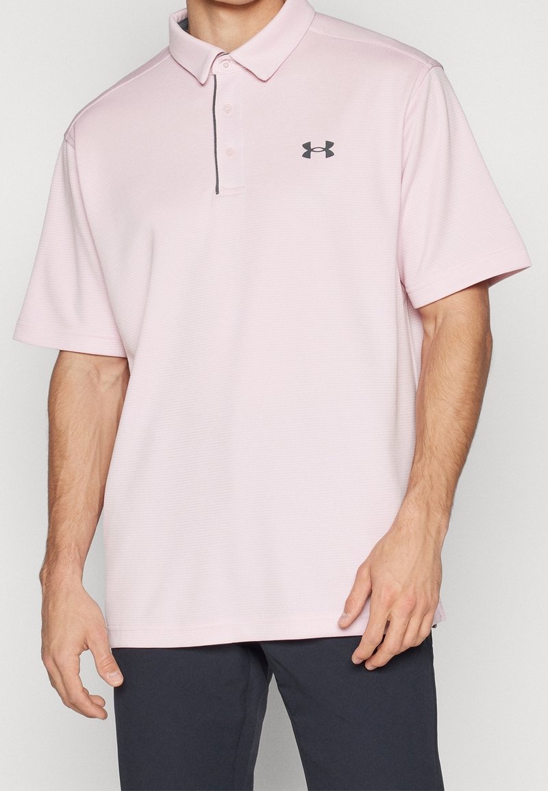 Homme portant un polo à manches courtes rose clair avec le logo Under Armour noir et un pantalon foncé, debout devant un fond uni.