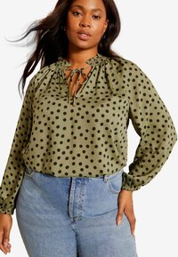 Olivegrüne Bluse mit Rüschensaum und Kordelzug. Mit schwarzen Punkten, langen Ärmeln und lockerem Schnitt. Kombiniert mit hoch geschnittenen Jeans.