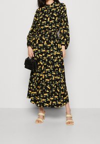 Donna che indossa un abito midi nero a maniche lunghe con stampa di frutta gialla, sandali con tacco beige e tiene in mano una piccola borsa nera.