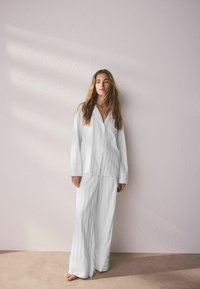 Femme aux longs cheveux debout pieds nus contre un mur clair, portant un haut de pyjama blanc ample boutonné et un pantalon à jambes larges.