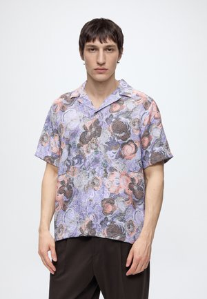 Bărbat purtând o cămașă cu mâneci scurte, nasturi, cu un model floral abstract în nuanțe de mov, roz și maro și pantaloni închiși la culoare, pe un fundal simplu.