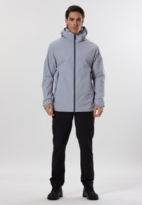 Veste imperméable gris clair avec un col haut, des zippers noirs et des manches ajustables. Associée à un pantalon noir et des chaussures noires.