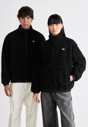 Twee modellen dragen zwarte fleece jassen met ritssluitingen aan de voorkant en Tommy Jeans logo's, staande tegen een effen witte achtergrond.