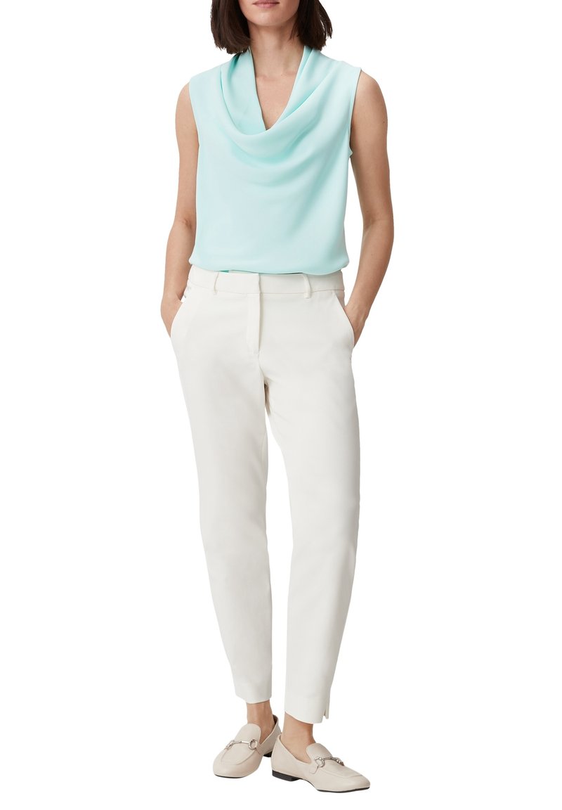 Femme portant un haut sans manches bleu clair drapé, un pantalon blanc ajusté et des mocassins beiges, debout les mains dans les poches.