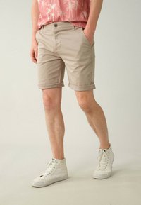 Shorts en coton beige avec revers, dotés de poches latérales et d'une fermeture à bouton à l'avant, associés à des baskets blanches montantes.