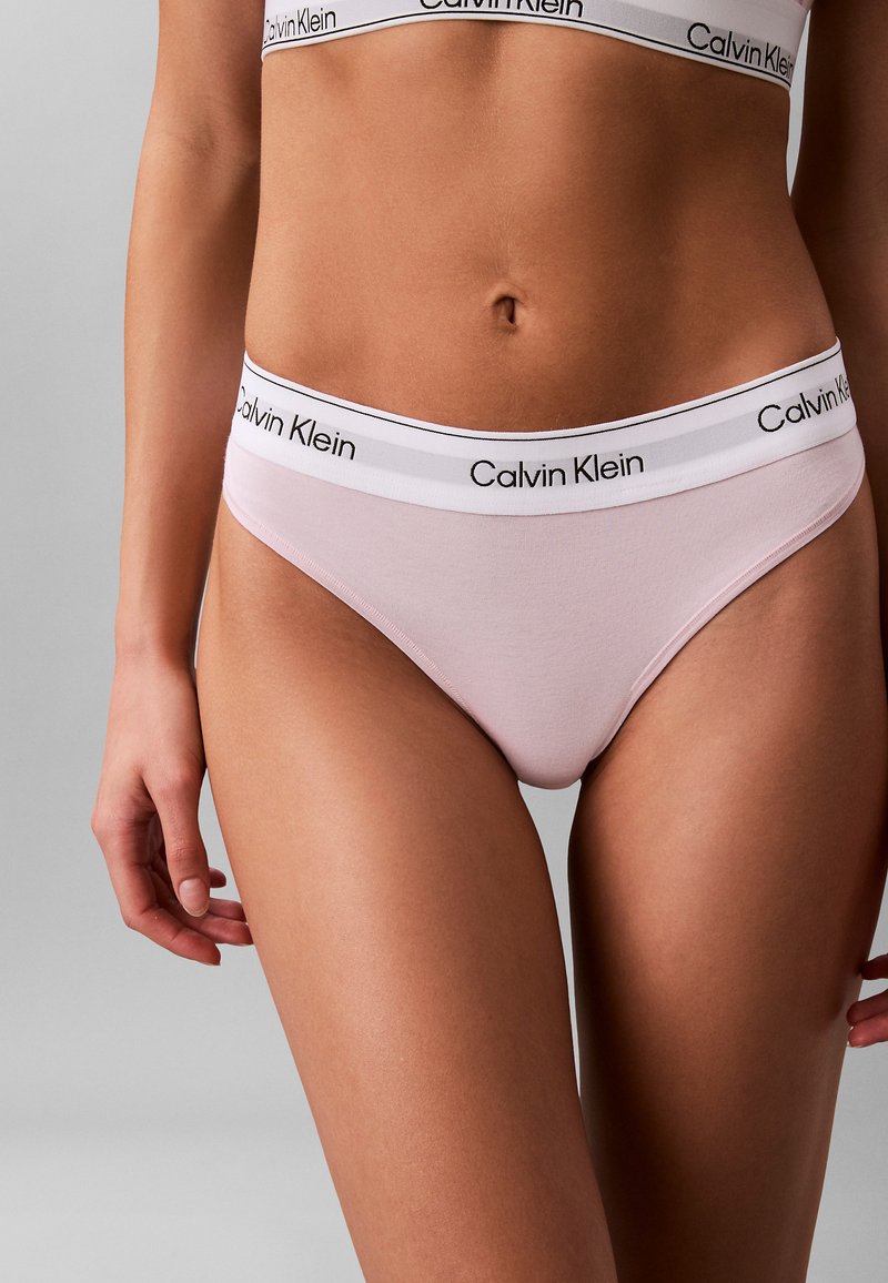 Vrouw draagt lichtroze Calvin Klein ondergoed met wit elastiek en merkopdruk, zichtbaar van de taille tot de bovenbenen.