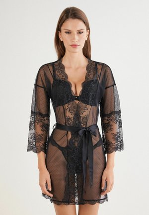 Robe en dentelle noire transparente avec un motif à pois, décolleté en V, manches trois-quarts et une ceinture en satin nouée à la taille.