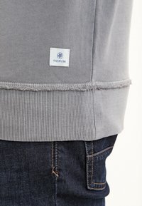 Ourlet côtelé gris de sweat-shirt avec une petite étiquette blanche « DENIM » au-dessus d'une couture à bords bruts, associé à une poche de jean denim bleu foncé et des surpiqûres orange.