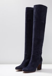 Stivaletti in camoscio blu navy, alti fino al ginocchio, con una punta leggermente arrotondata e un tacco spesso. La texture è morbida, con un aspetto liscio e uniforme.