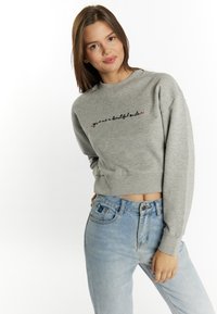 Grijze cropped sweatshirt met zwart en roze geborduurde tekst "geef me een mooie glimlach" met lange mouwen en geribde manchetten. Gemaakt met lichtblauwe jeans.