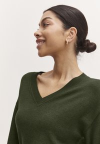 Maglione verde a maglia con scollatura a V, texture morbida e dettagli a coste. La modella ha un'acconciatura a bun e orecchini a cerchio minimalisti in oro.