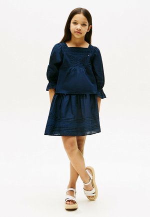 Ragazza che indossa un abito blu scuro con scollo quadrato e maniche a sbuffo, in piedi con le gambe incrociate, indossa sandali bianchi con cinturini alla caviglia.