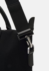 Sac en toile noir avec une large sangle noire et un fermoir métallique à finition argentée. Des accents en cuir améliorent la durabilité et la structure.