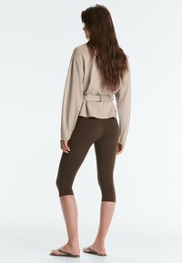 Femme debout, de dos, vêtue d’un haut beige ceinturé à manches longues, de leggings courts marron foncé et de sandales plates sur fond blanc.