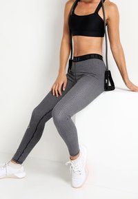 Musta urheiluliivit ja kuvioidut musta-valkoiset legginsit, joissa on korkea vyötärö. Malli istuu seinää vasten valkoisissa urheilu kengissä.