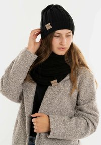 Zwarte gebreide beanie en sjaal met een gestructureerd patroon. De beanie heeft een omgeslagen rand; beide hebben een klein stoffen label. Lichtgrijze fuzzy jas.