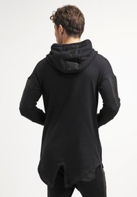Sudadera negra con capucha, con detalles acanalados en las mangas y la parte trasera. Corte largo con un dobladillo característico en forma de split. Textura suave.