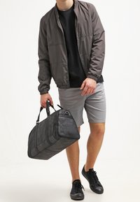 Zwart leren sporttas met dubbele handvatten, rechthoekige vorm, gestructureerd uiterlijk en een ritszak; gedragen met een grijze bomberjack en shorts.