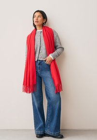 Écharpe rouge à franges drapée sur un cardigan gris à boutons, associée à un jean évasé bleu et des chaussures noires. Textures lisses et couleurs vives.