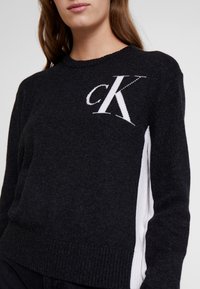 Svart stickad tröja med vita accenter, med rundad halsringning och en framträdande broderad 'CK'-logotyp på framsidan.