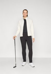 Weißer Reißverschlussjacke mit strukturiertem Muster, schwarzes Poloshirt, schwarze Leggings und weiße Sportschuhe; hält einen Golfschläger.