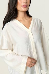 White, loose-fitting blouse with a V-neckline, long sleeves, and button closure. Soft, lightweight fabric with a subtle texture.