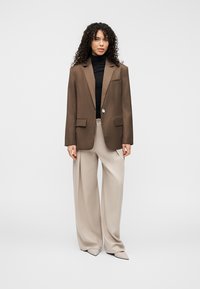 Blazer marron sur mesure avec un bouton unique, poches avant, porté sur un col roulé noir, associé à un pantalon à jambes larges beige et des chaussures pointues.