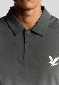 Polo di colore grigio scuro con una texture morbida, dotato di un colletto a tre bottoni e un logo di un'aquila bianca sulla parte sinistra del petto.