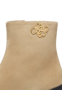 Stivale alla caviglia in suede beige con una texture liscia, caratterizzato da un emblema decorativo dorato sul lato. Design semplice e bordo superiore arrotondato.