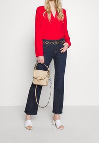 Blouse rouge à manches longues avec un décolleté en V, jeans foncés avec une ceinture en chaîne dorée, un sac à main texturé beige et des sandales à talons blocs blanches.