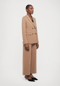 Dvojřadý blazer a široké kalhoty z jemného hnědého materiálu. Úzké klopy, boční kapsy a černé vysoké podpatky dotvářejí celkový vzhled.