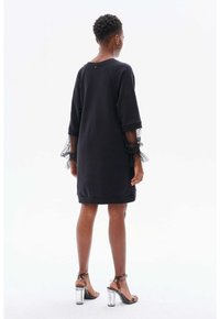 Schwarzes Sweatshirtkleid mit dreiviertellangen Tüllärmeln, umgeschlagenem Saum und zartem Knopfdetail am Rücken. Kombiniert mit transparenten, hohen Sandalen.