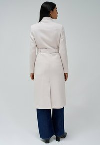 Cappotto lungo di un beige chiaro con colletto strutturato, vita con cintura e silhouette dritta. Abbinato a jeans a gamba larga blu scuro e stivali neri alla caviglia.