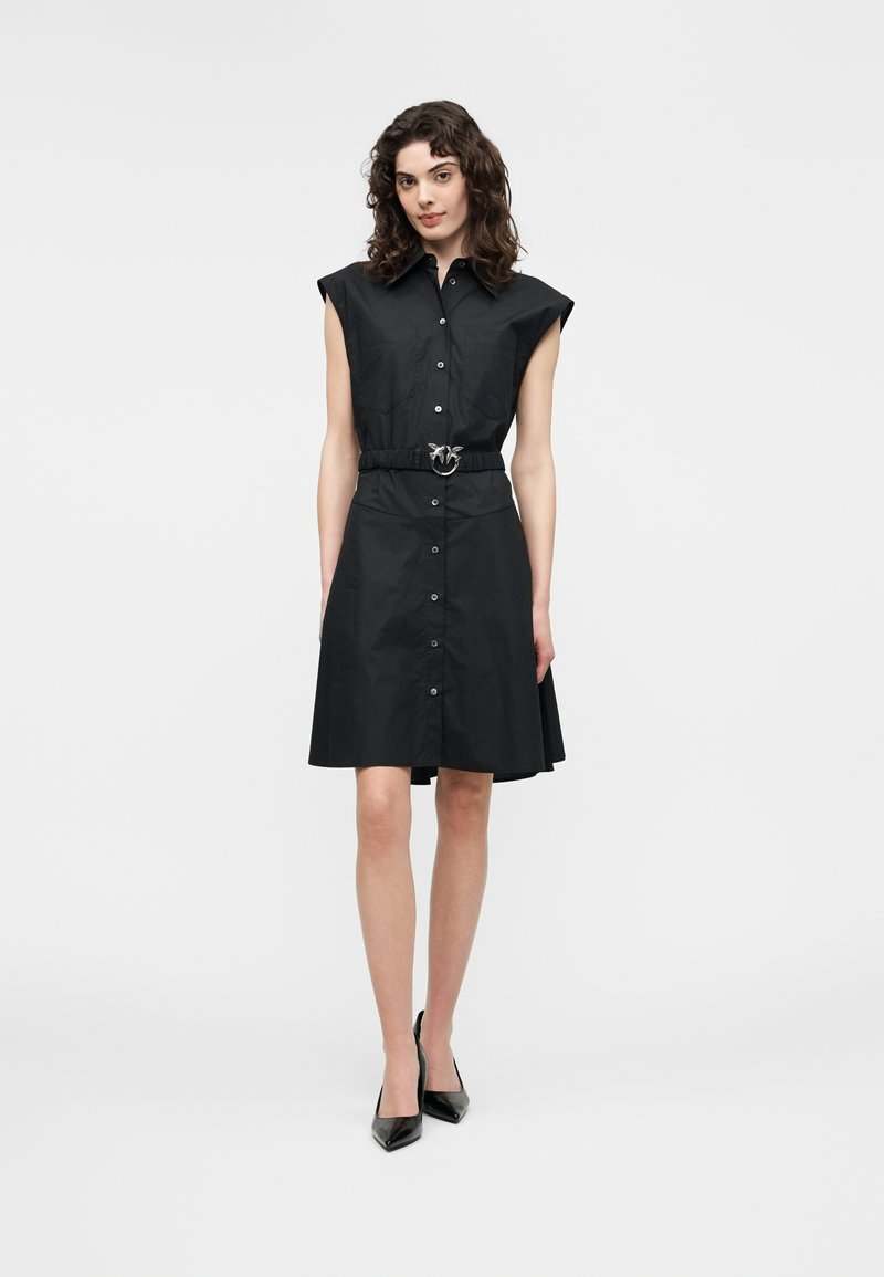 Γυναίκα με σγουρά μαλλιά, φοράει μαύρο αμάνικο φόρεμα με κουμπιά και ζώνη, καθώς και μαύρα παπούτσια με τακούνι, στέκεται μπροστά σε απλό λευκό φόντο.